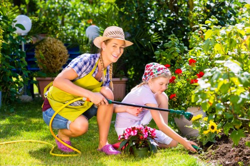 A woman and a young girl are gardening together in a lush, well-maintained outdoor space that appears to be part of a private garden or backyard in Lee. The woman, wearing a straw sunhat, a blue and white checkered shirt, and a bright yellow apron, is kneeling on the grass and smiling as she holds a yellow garden hose. The girl, dressed in a light purple outfit with a floral-patterned sun hat, is actively planting or tending to flower beds that include vibrant red, yellow, and pink blooms. The garden features a neatly trimmed lawn with dense, leafy green bushes and trees in the background, along with a variety of flowering plants and decorative garden elements such as hanging lanterns or lights. Sunlight filters through the foliage, suggesting a warm, sunny day, creating a cheerful and productive outdoor environment suitable for gardening and family outdoor activities. The scene reflects typical garden maintenance activities, supported by gardening services in Lee to maintain the landscape's tidy and vibrant appearance.