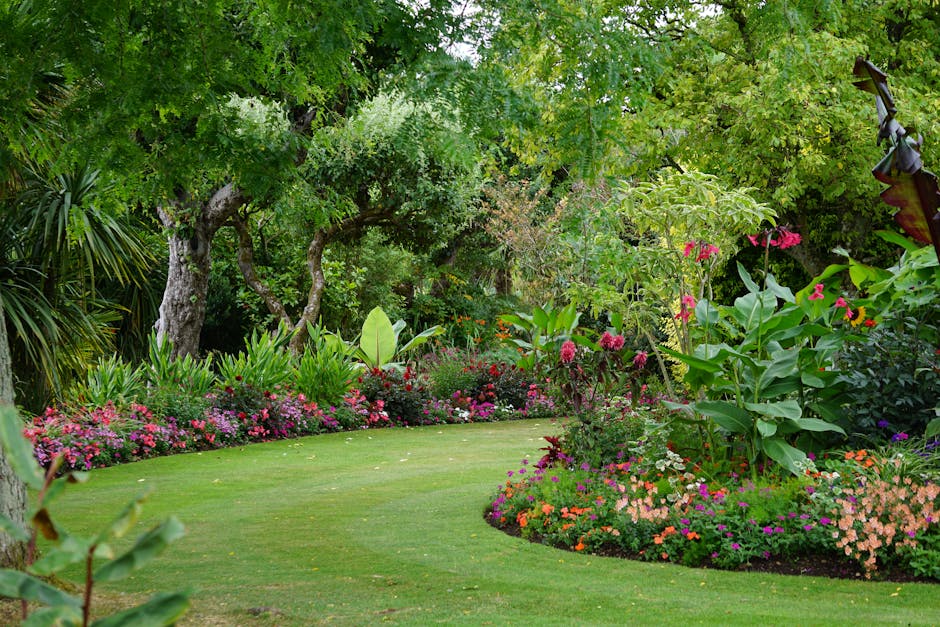 A meticulously maintained garden with a lush, green lawn bordered by vibrant flower beds filled with a variety of colorful blooms, including pink, purple, and orange flowers. Mature trees with texture